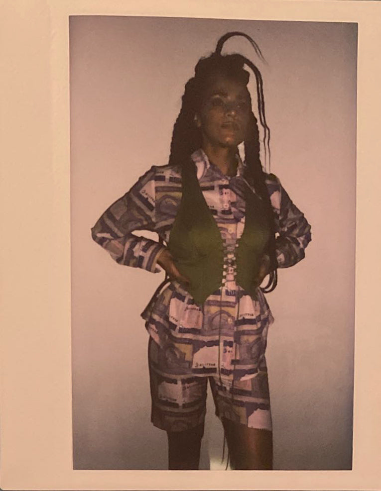 A polaroid of a woman standing in 100 Afghani bill printed shorts and long sleeve shirt, with a green corset on top.