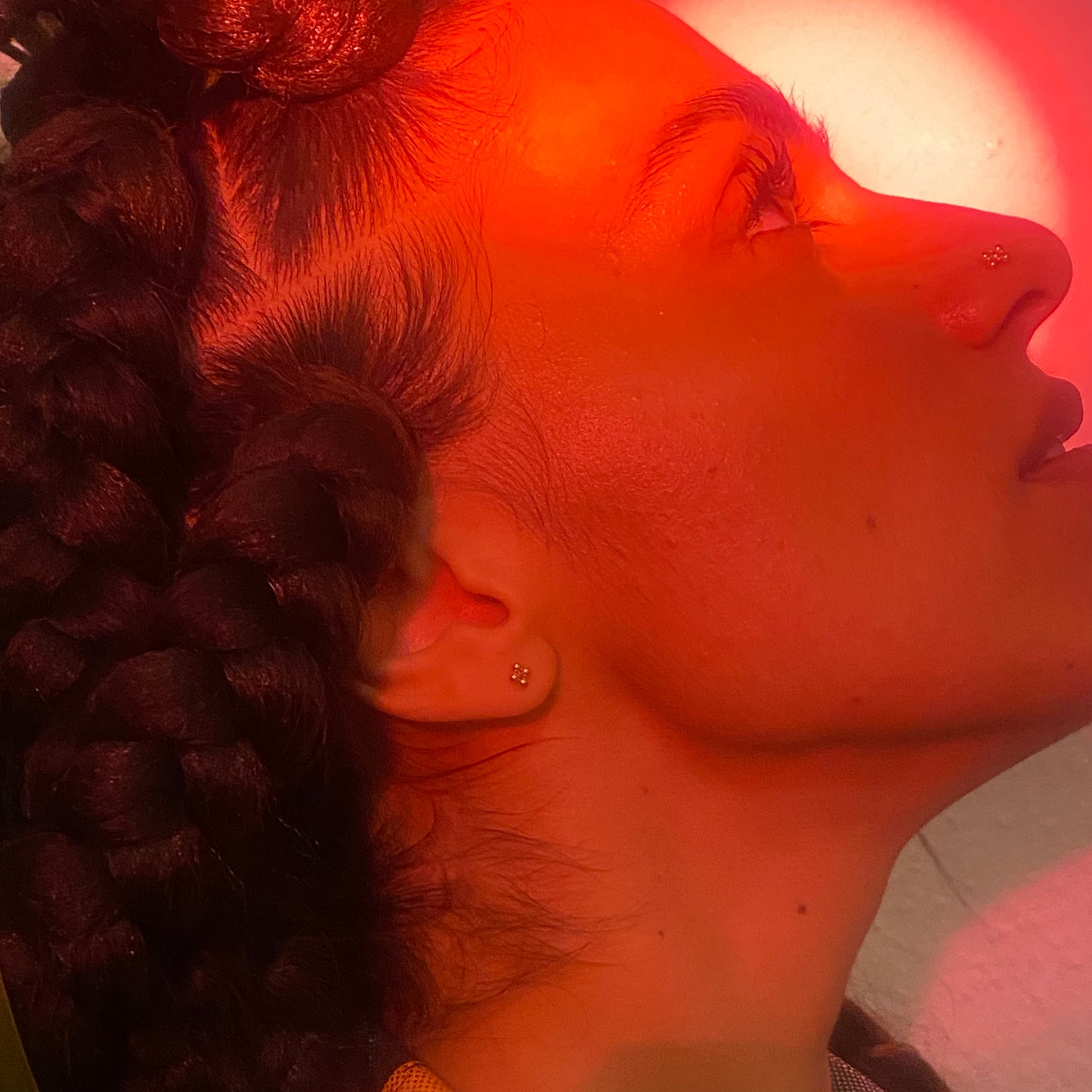 A woman lit up by a red light looks upwards, and her gold nose stud and ear stud shine in the light.