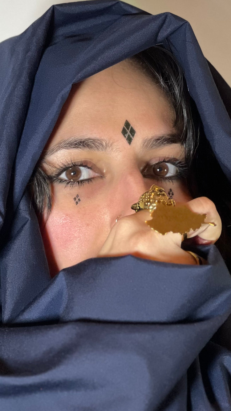 Photo of a woman with tribal tattoos in diamond formation on her cheeks and forehead, her face covered by her hand. 