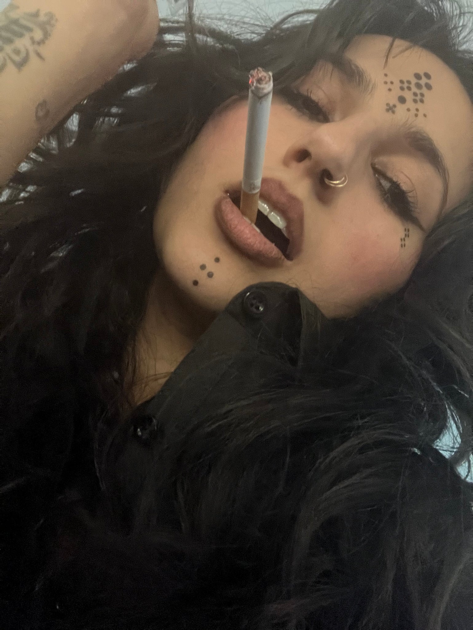 Photo of a woman with tribal tattoos in diamond formation on her cheeks, forehead and chin in intricate patterns with a cigarette in her mouth. 