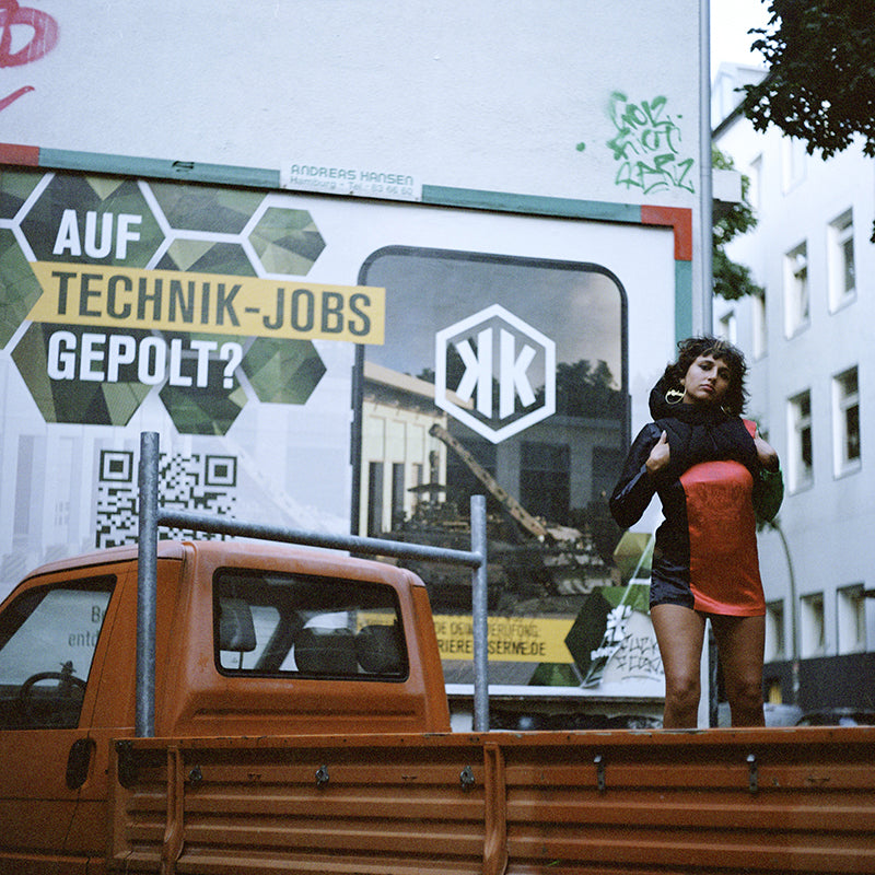A woman in dress that is the colour of the Afghan flag, and a black puffy vest, in front of a German army recruitment poster, on top of an orange truck.