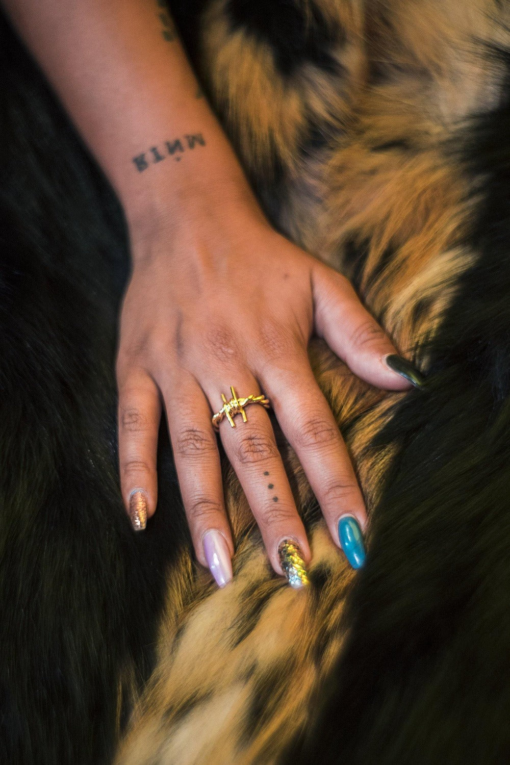 The Kabul House Essential Barb Wire Ring - Blingistan, hand with tattoos wearing gold barbwire ring, on fur backdrop.