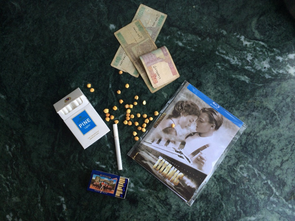 Afghani money, pine cigarettes, chickpea nuts, matches and a blue ray disk or Titanic on a green marble background.