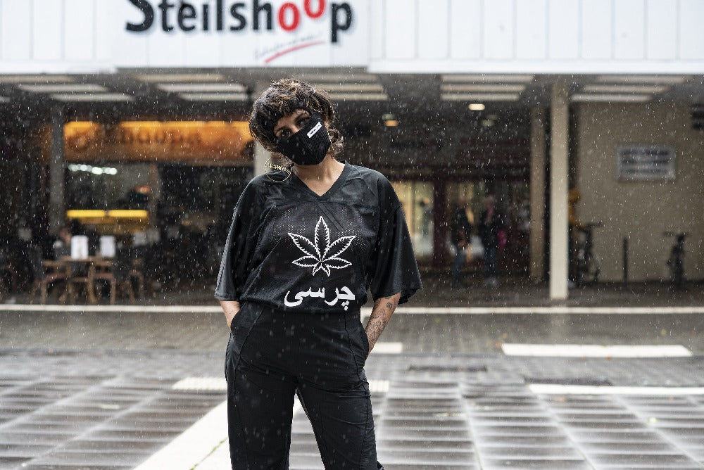 A woman in front of a building that reads "Steilshoop", in all black shirt, pants and balaklava. The shirt says "charsi" with a weed leaf in the middle. 
