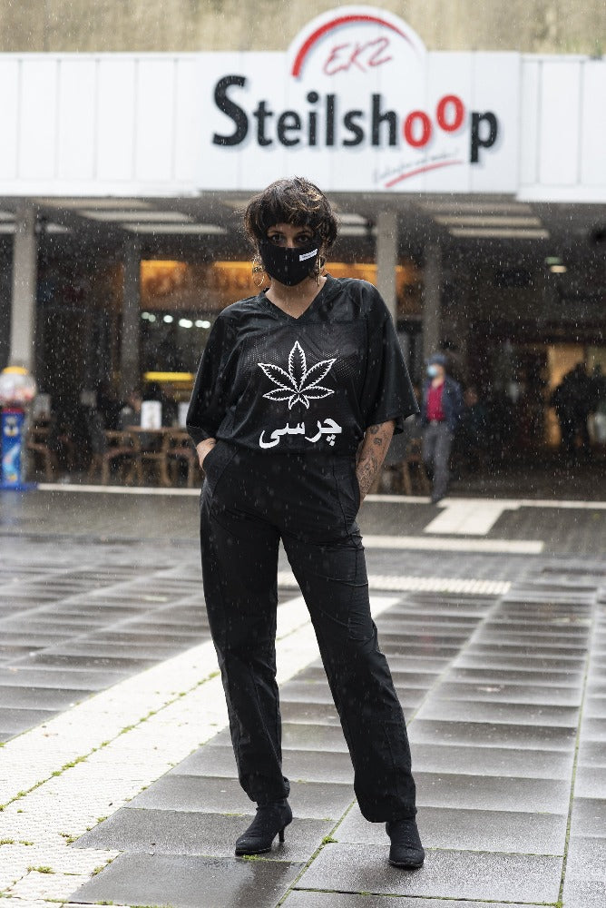 A woman in front of a building that reads "Steilshoop", in all black shirt, pants and balaklava. The shirt says "charsi" with a weed leaf in the middle. 