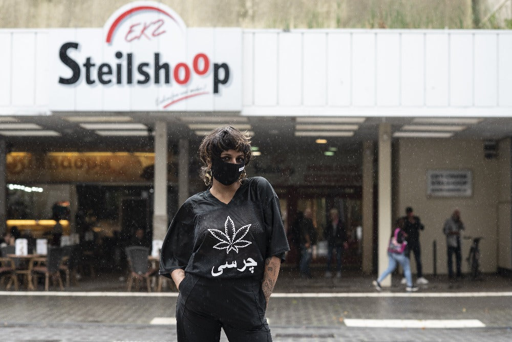 A woman in front of a building that reads "Steilshoop", in all black shirt, pants and balaklava. The shirt says "charsi" with a weed leaf in the middle. 