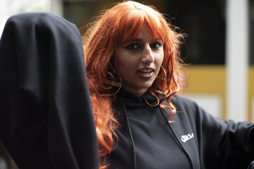 a woman with long orange hair, mouth slightly open, looking at the camera, with a black sweatshirt on and hoops that say "Lewanai".