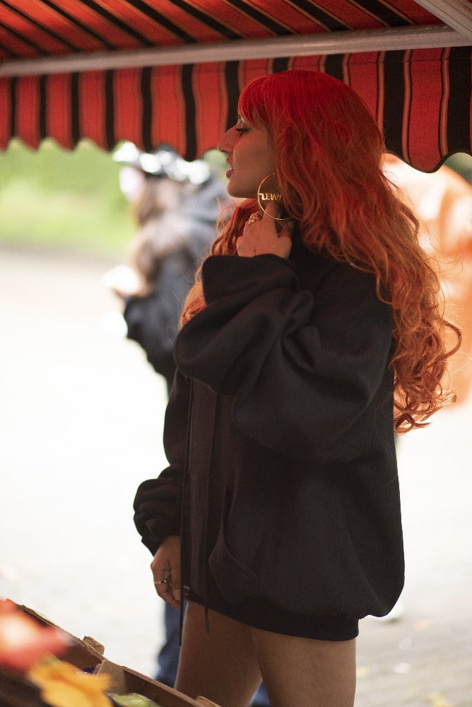 a woman with long orange hair, at a vegetable market, playing with a gold hoop in her ear, in a big black sweatshirt.