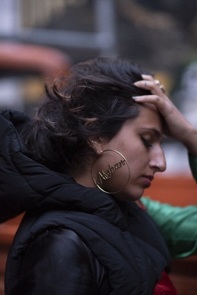 woman with eyes closed, side profile, with big gold hoops that say Afghan, with a black puffy jacket.