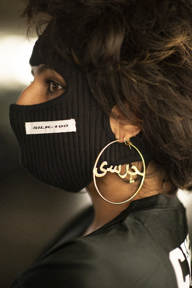A woman in a black balaklava with the word Silk-100 written on it. Woman wears gold hoops with the word "Charsi" in Dari.