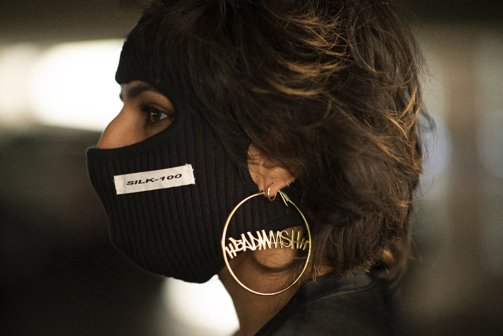 a woman turned away from the camera in a black balaklava with the word Silk-100 stitched on it, wearing gold hoops with the word "Badmaash" in the middle.