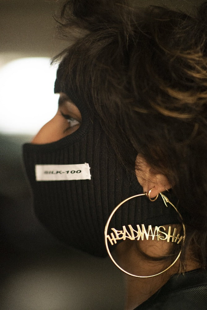 a woman turned away from the camera in a black balaklava with the word Silk-100 stitched on it, wearing gold hoops with the word "Badmaash" in the middle.