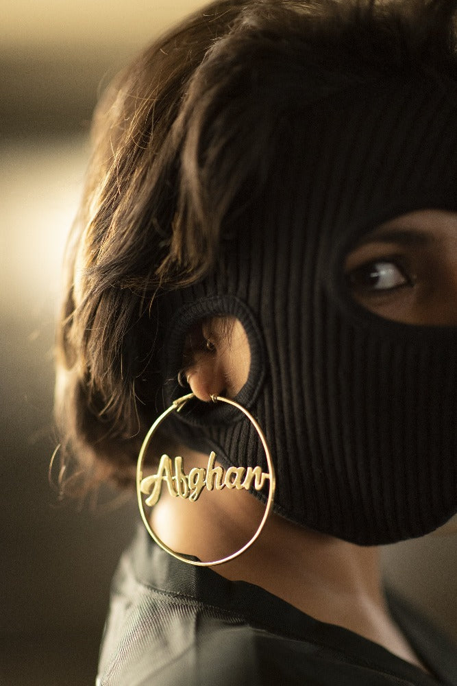 woman looking towards camera with a balaklava and gold hoops that say Afghan