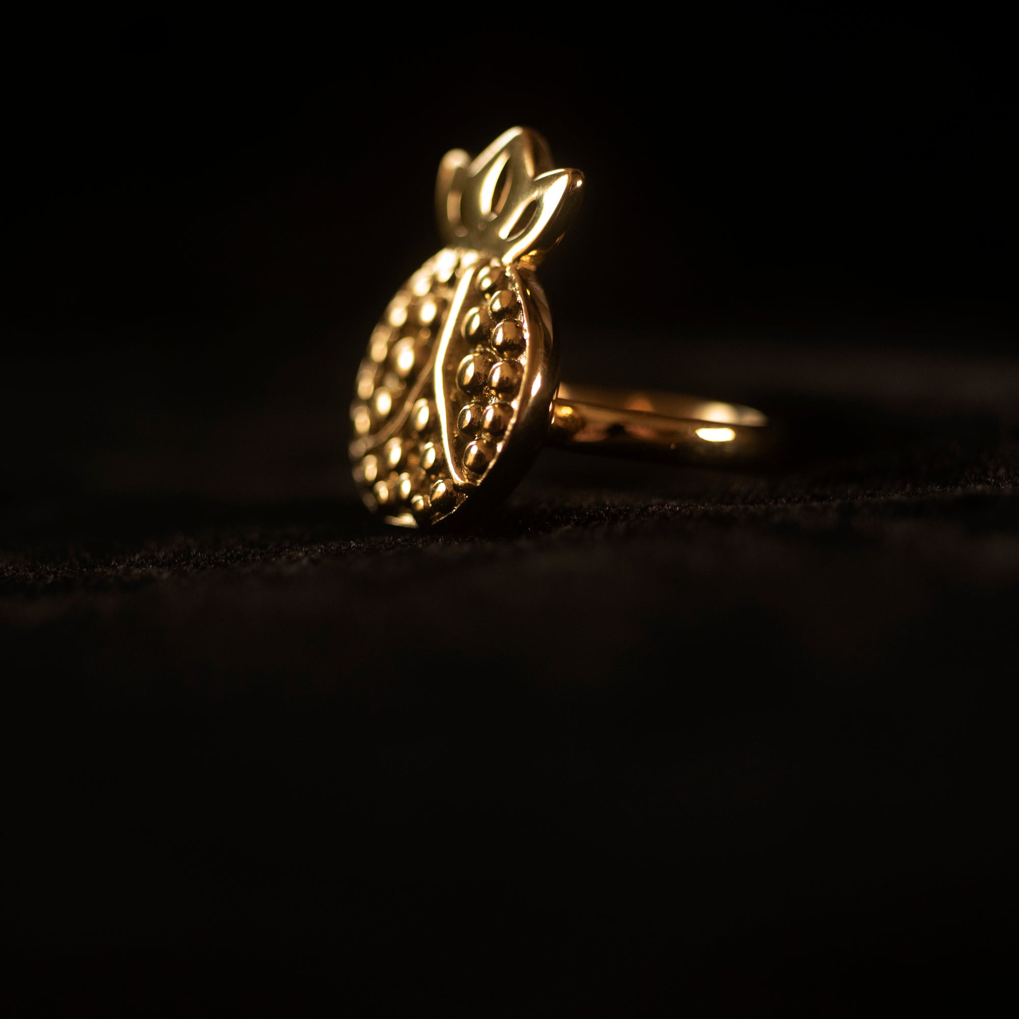 Against a black backdrop sits a pomegranate shaped gold ring, turned to the side. 