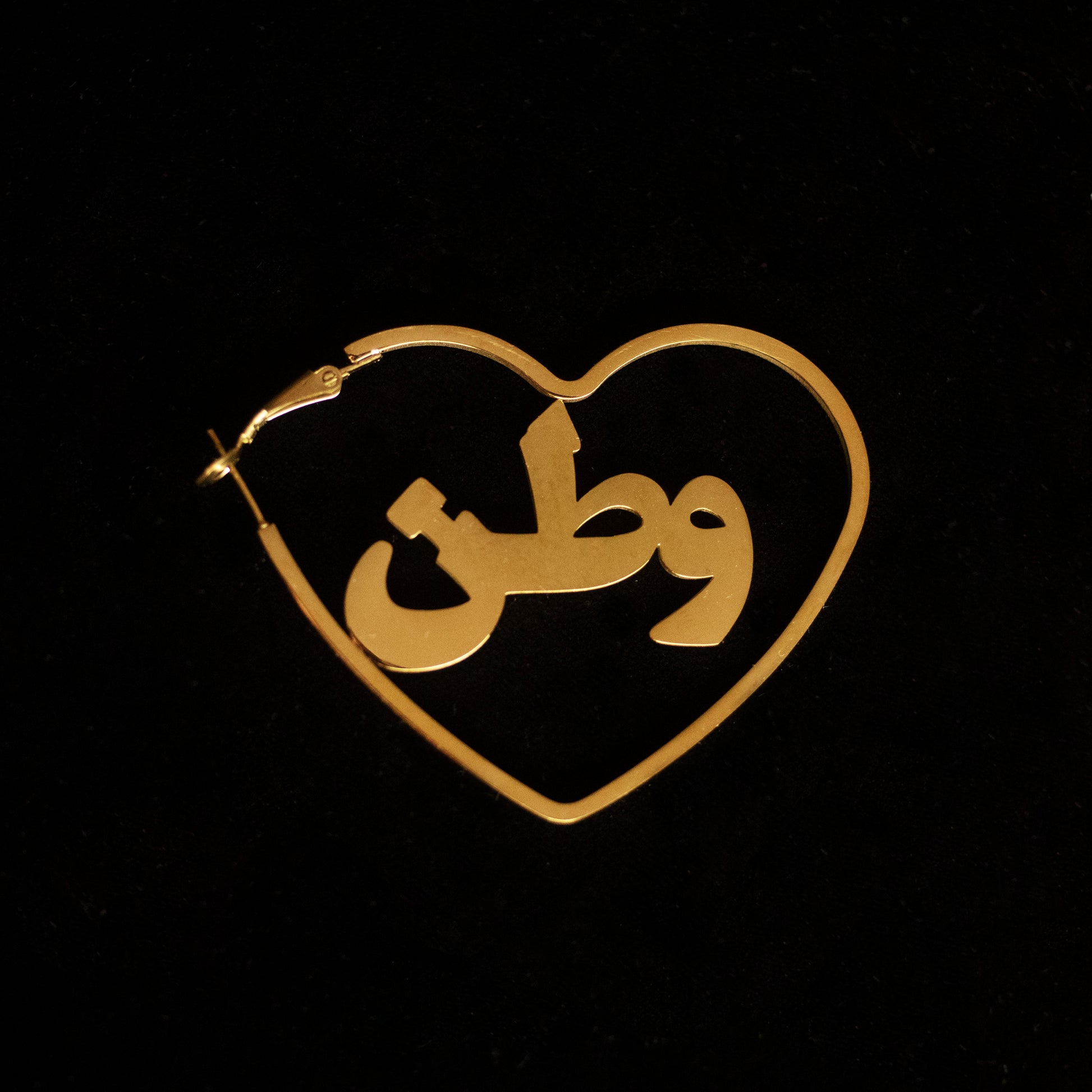 Gold hoops in the shape of a heart with the word 'watan' on it against a black back drop.
