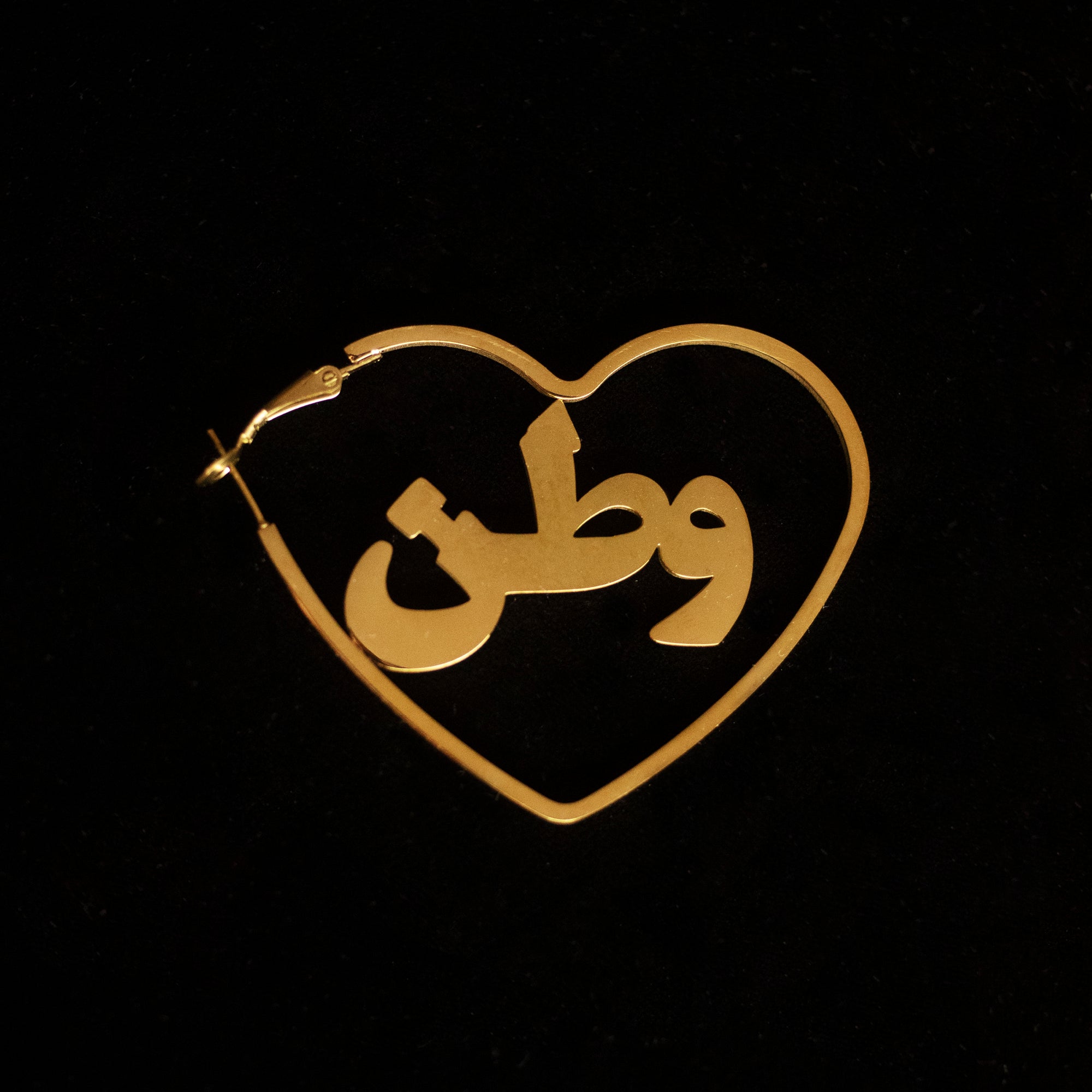 Gold hoops in the shape of a heart with the word 'watan' on it against a black back drop.