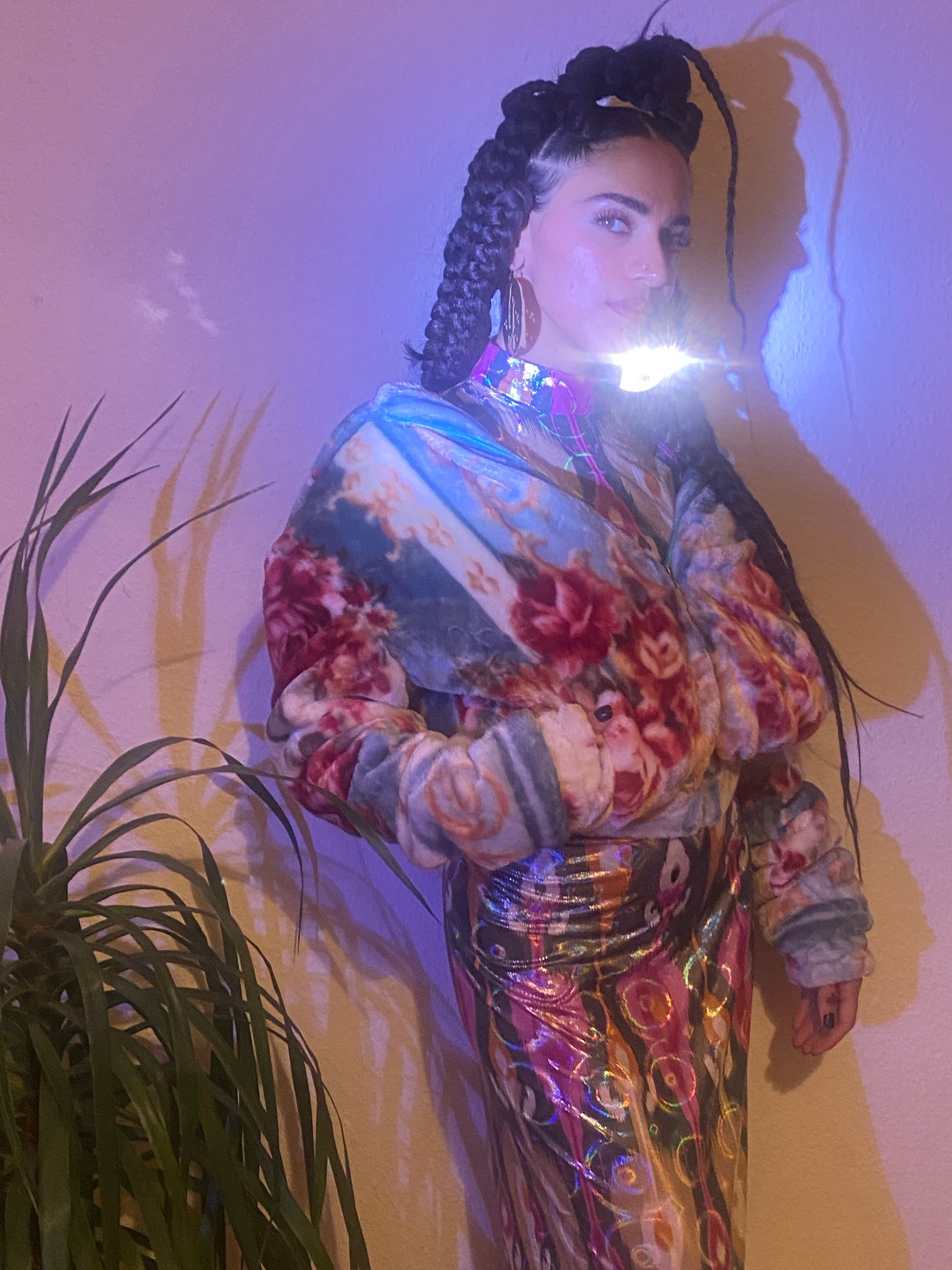 Woman with braids next to a plant wearing a shiny dress and a blanket jacket.