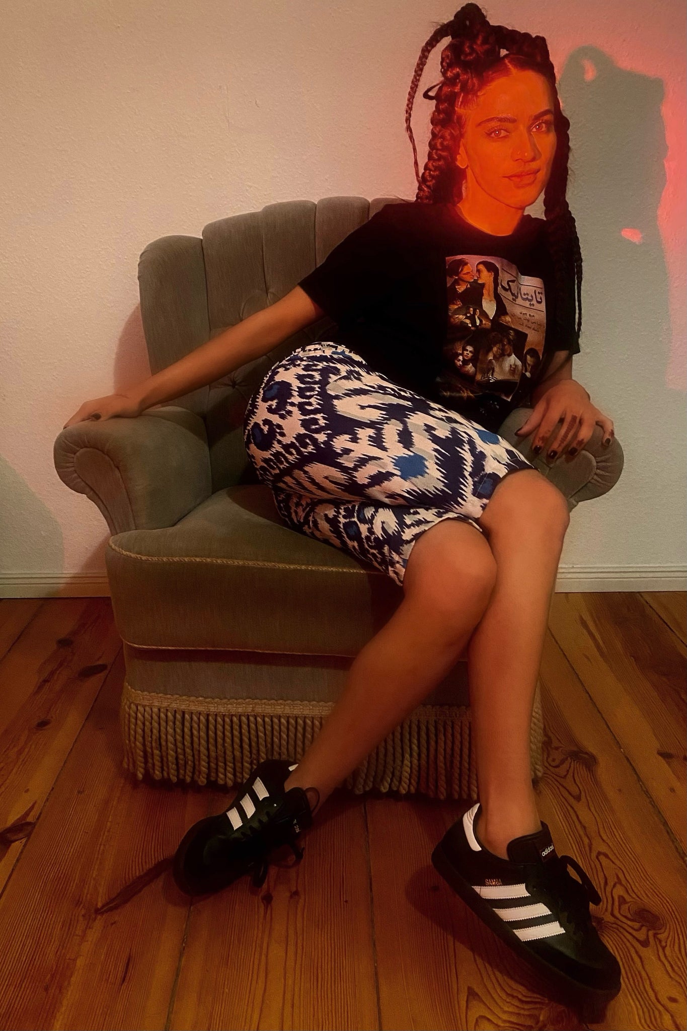 A red light shines on a woman on a grey chair, wearing Adidas Sambas, a Titanic graphic tee, and adras/ikat capris