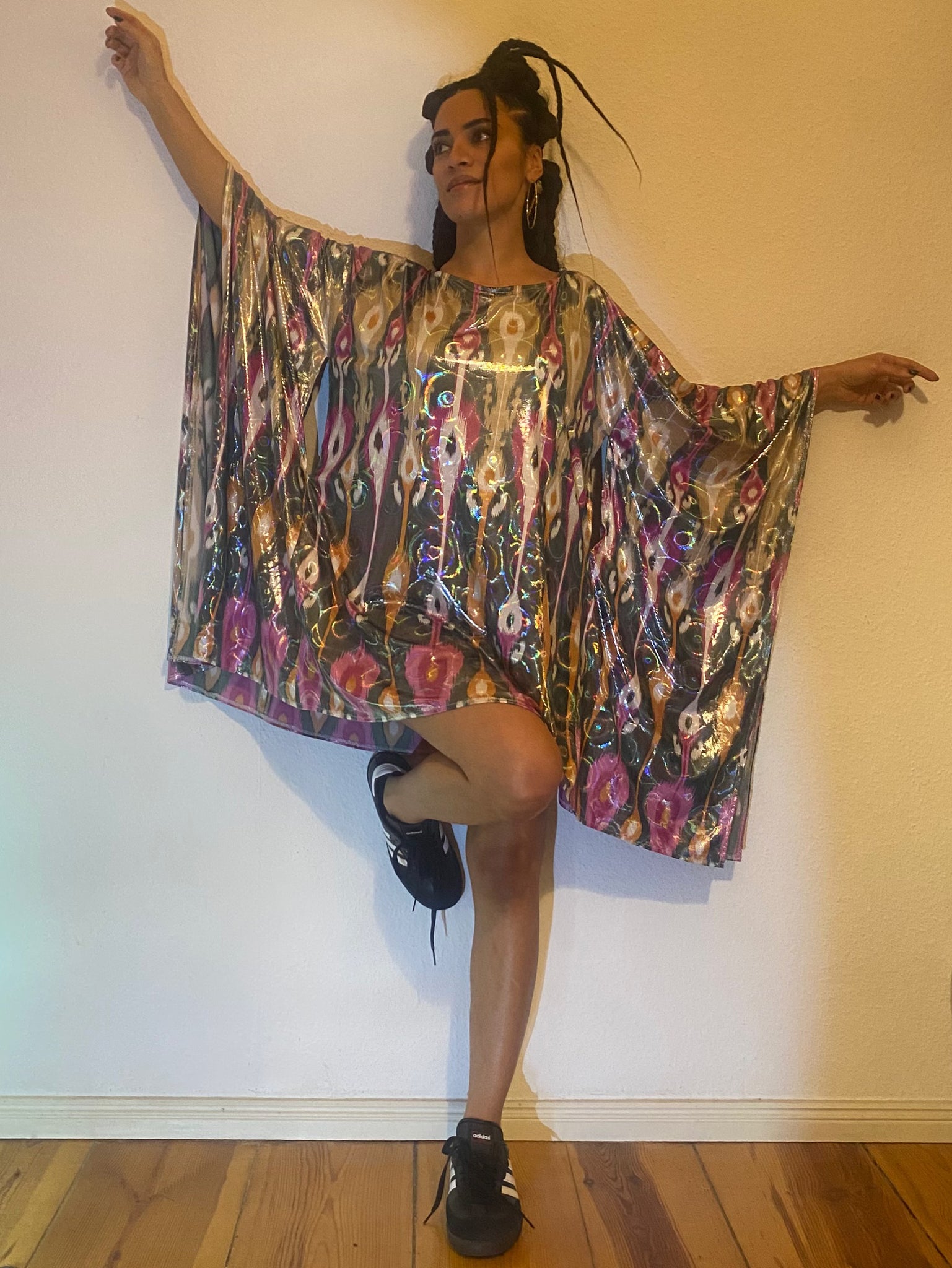 A woman poses against a white wall with a colourful dress, her arms up and one of her legs up.