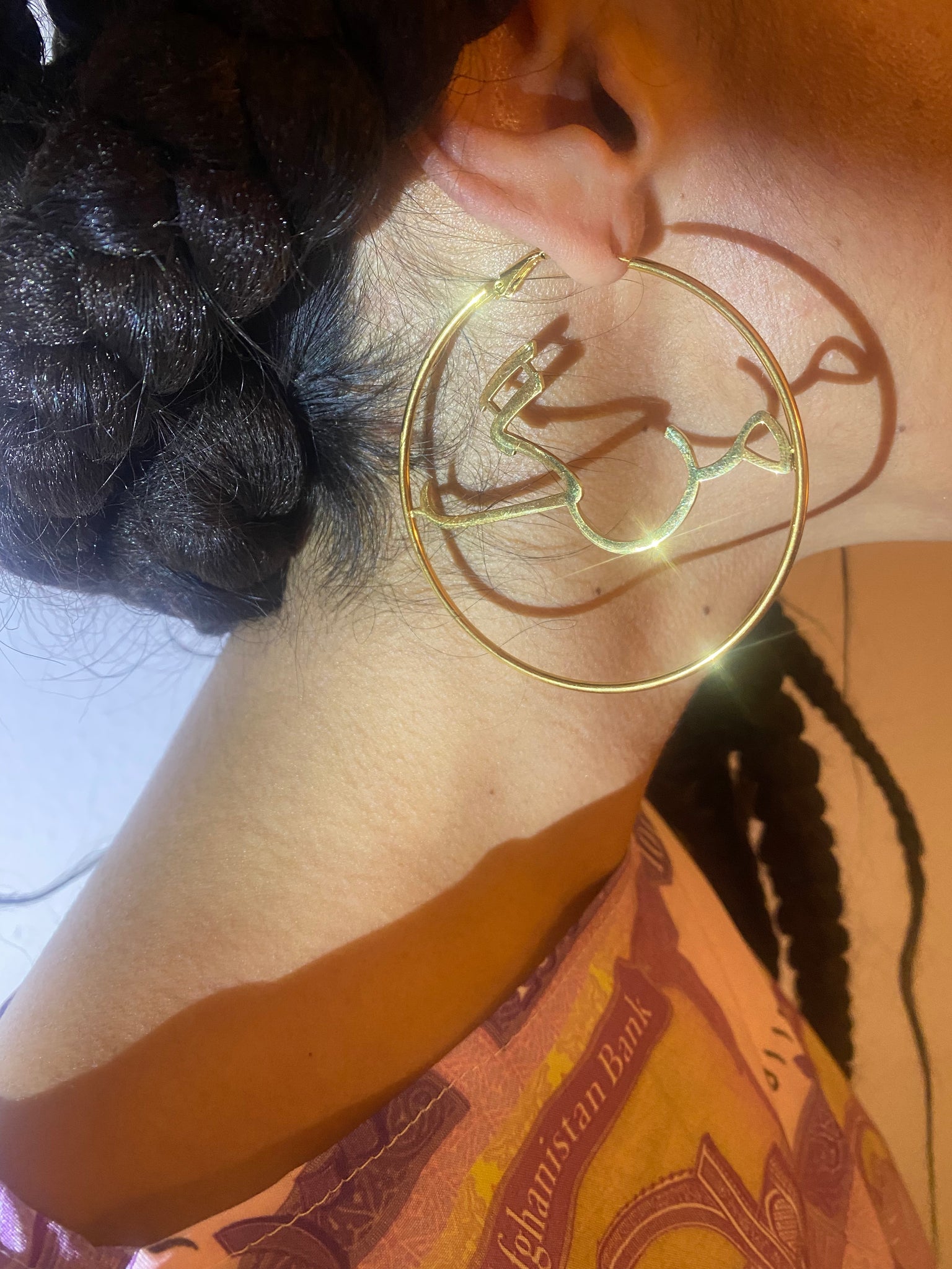 A close up of a woman's ear with sparkling gold hoop earrings with the word 'marg' in Dari on the inside.