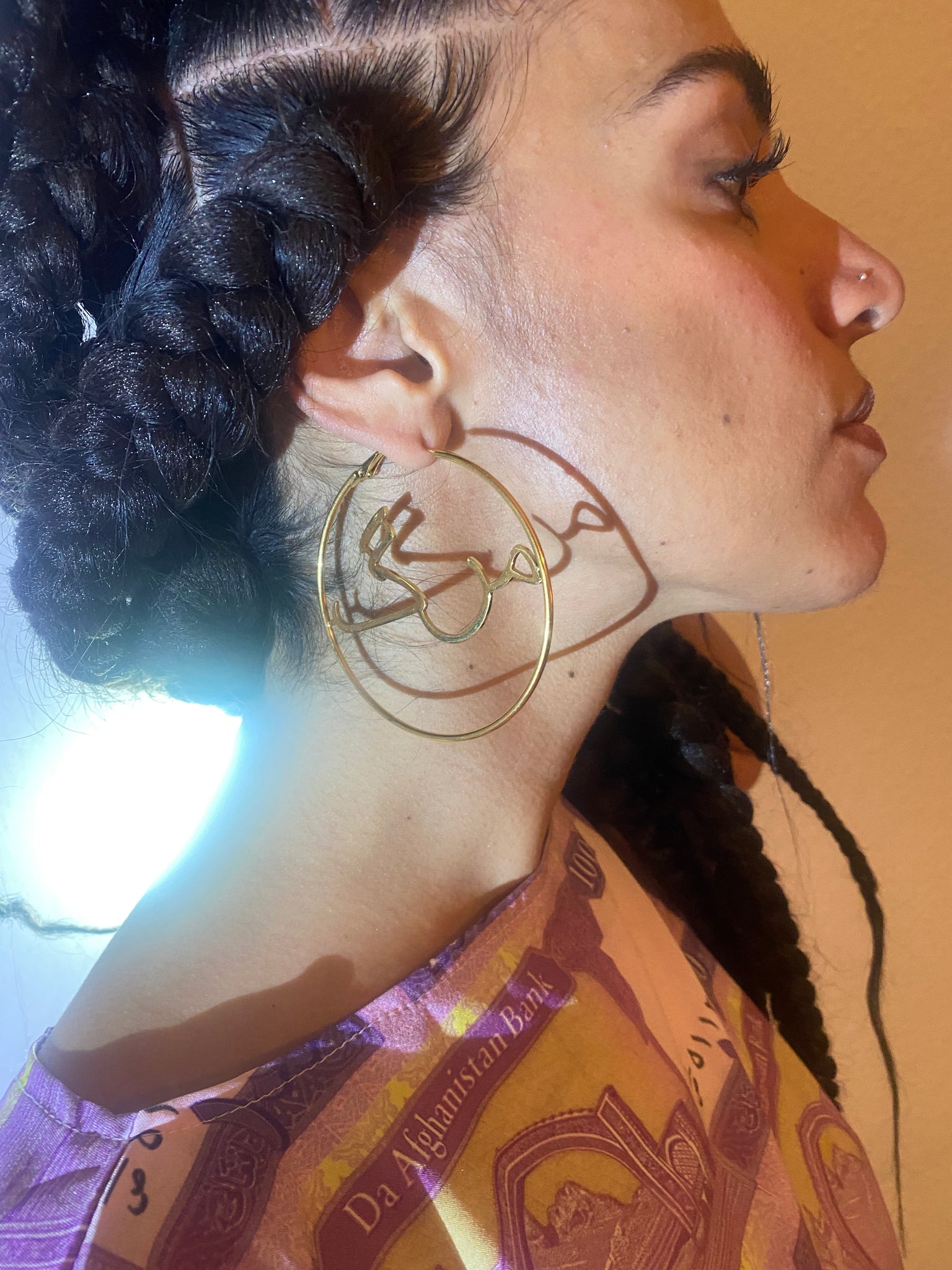 A woman with braids profile while she wears gold hoops with the word 'marg' in the middle in Dari.