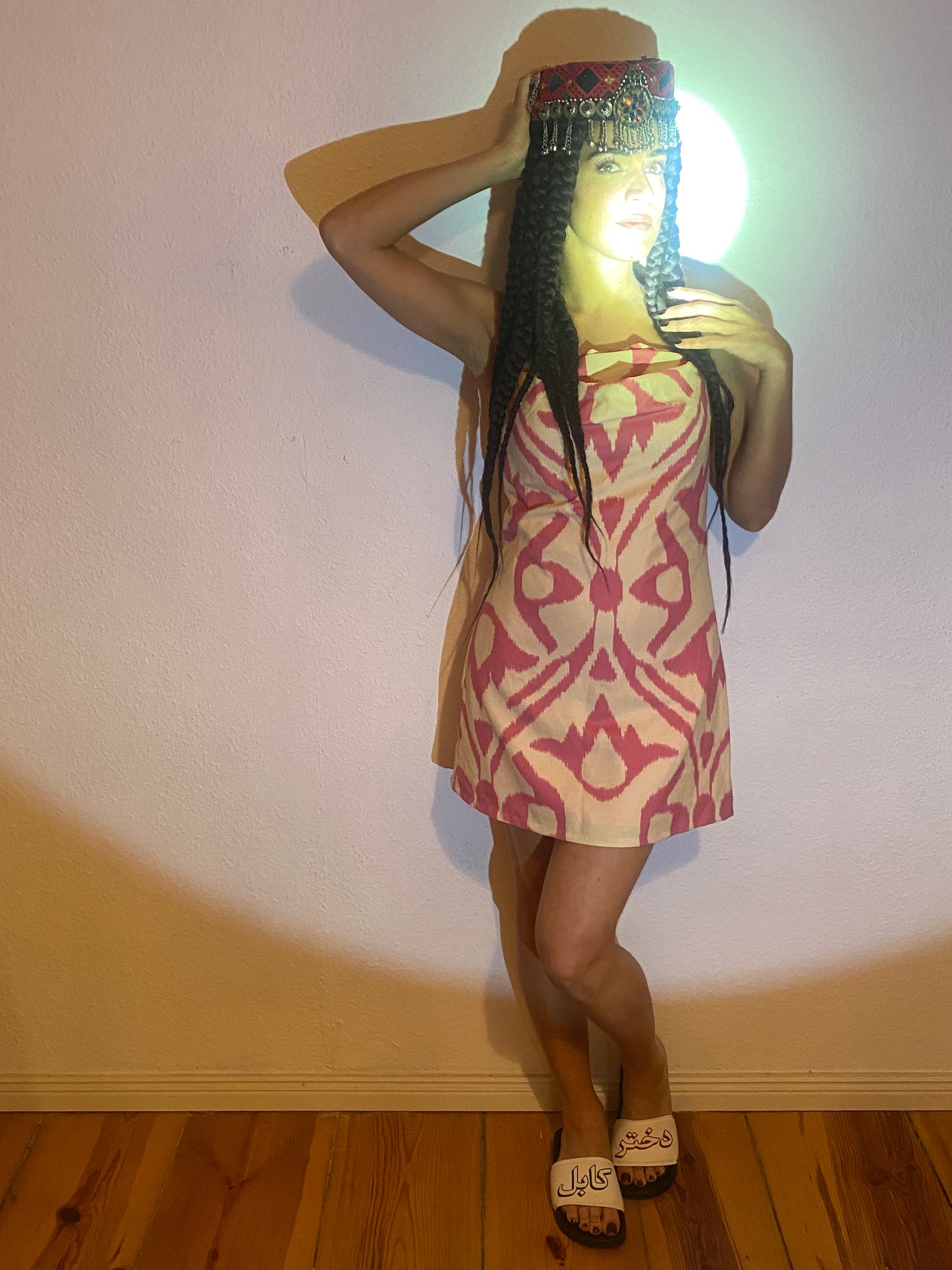 Women standing against a wall in a pink and cream dress, light shining on to her face.