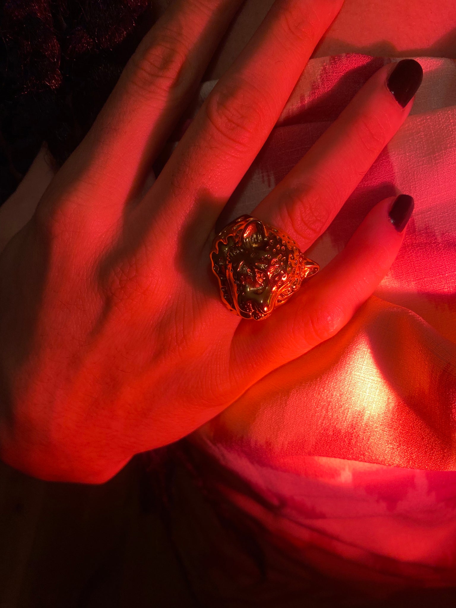 With moody red lighting a woman with painted nails wears a ring shaped as a snow leopard's head. 