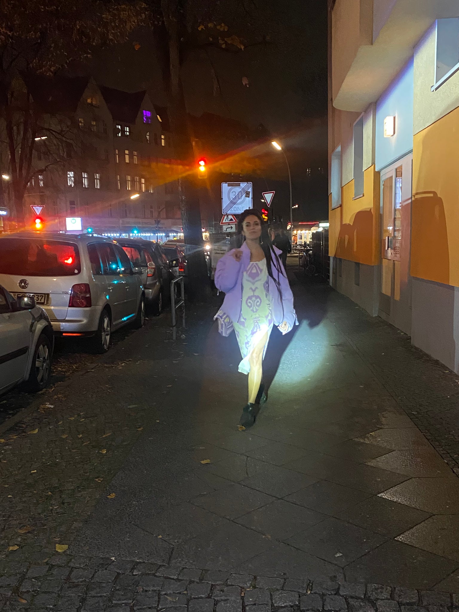 A woman in white and lavender walks a Berlin city street at night.