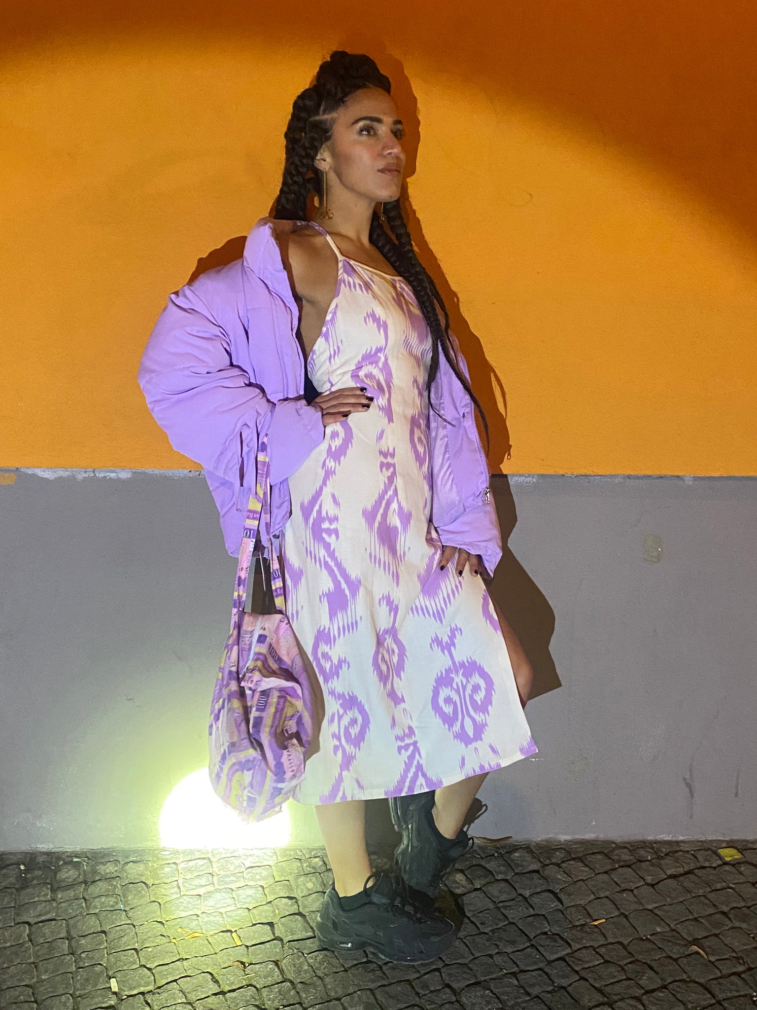A woman strikes a pose on a night street showing the curve of her back in a white and lavender adras dress against an orange and grey wall.