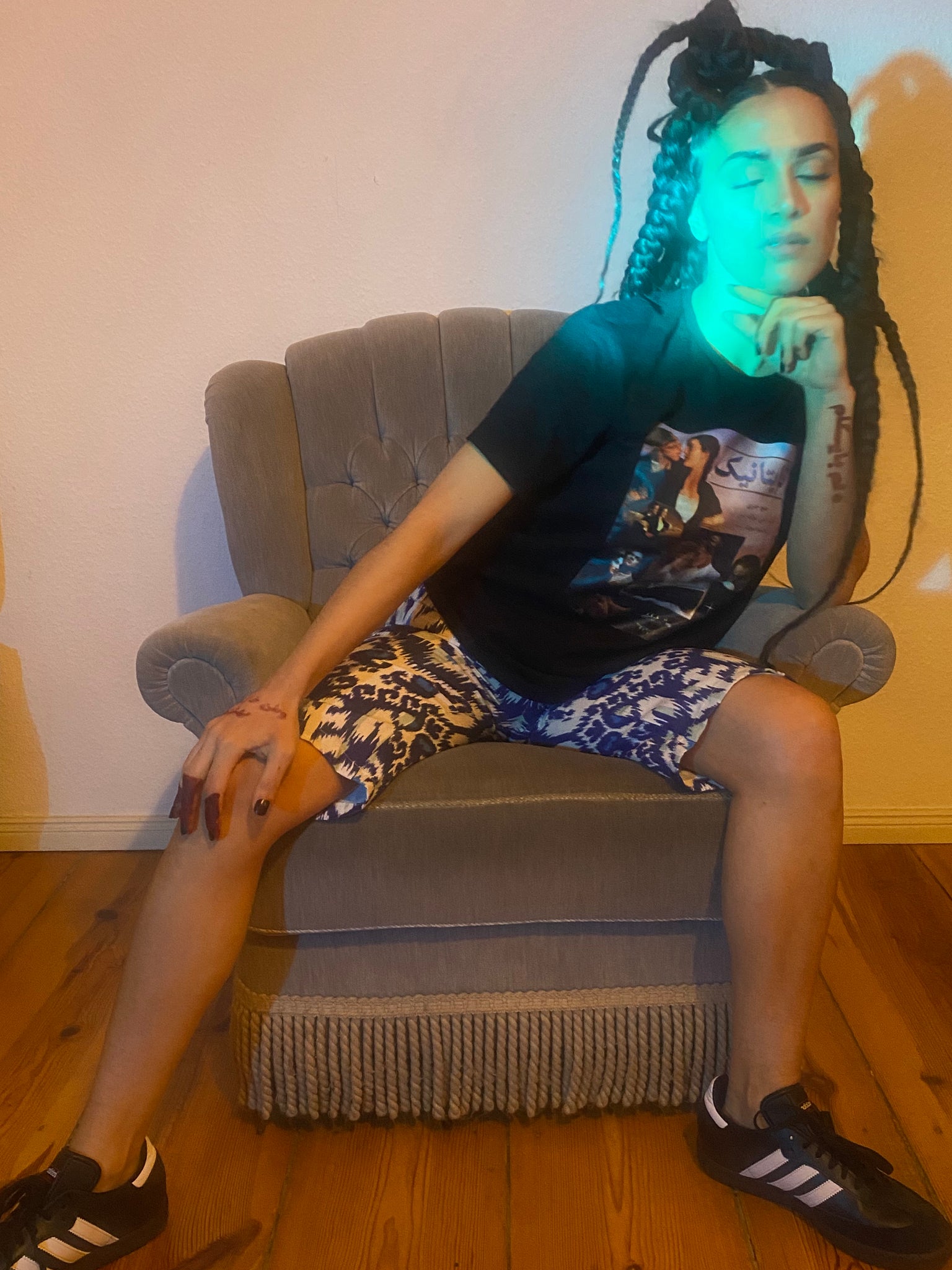 A blue green light shines on a woman whose eyes are closed on a grey chair, wearing Adidas Sambas, a Titanic graphic tee, and adras/ikat capris
