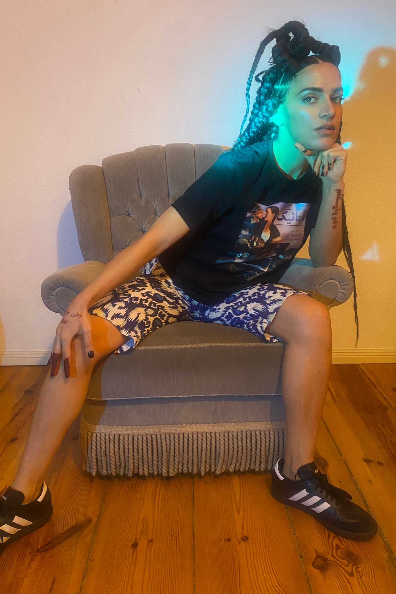 A green light shines on a woman on a grey chair, wearing Adidas Sambas, a Titanic graphic tee, and adras/ikat capris
