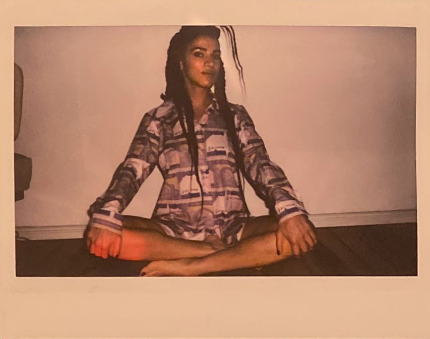 A polaroid of a woman sitting cross legged with long braids and a purple outfit.