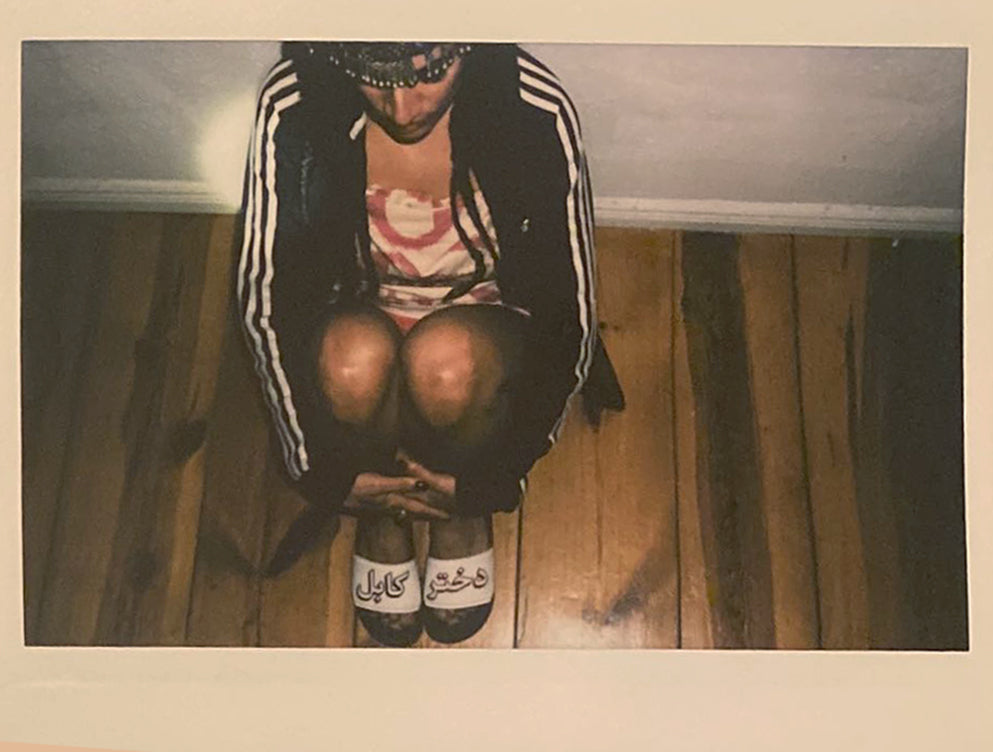 Polaroid of a woman sitting on the ground, her sandals read "Kabul Girl" in Dari, wearing an Adidas tracksuit and an Uzbeki adras mini dress.