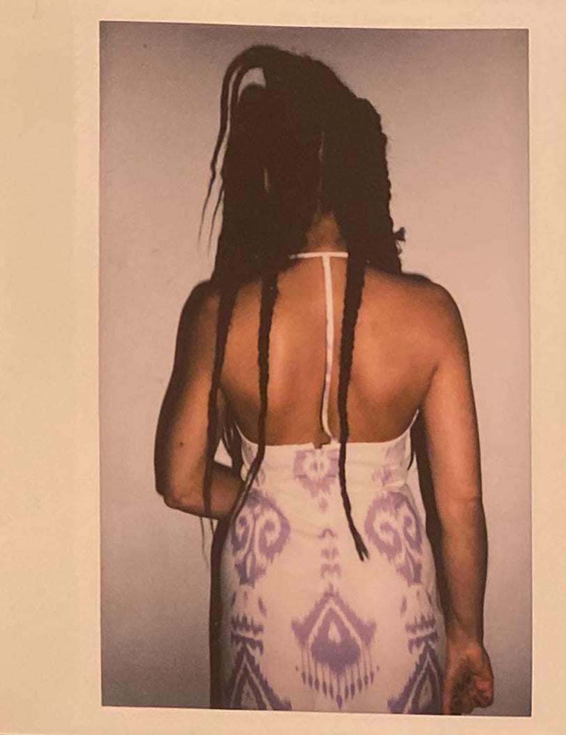 A polaroid of a woman with braids back wearing a white and lavender adras dress.