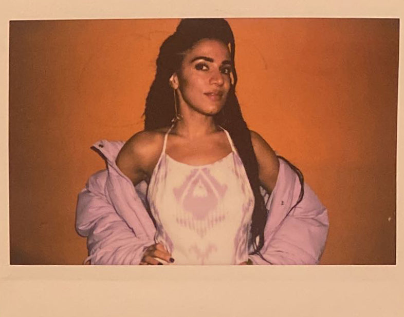 A polaroid of a woman smiling to the camera with an orange background, she wears a lavendar jacket and a white and lavendar adras/ikat dress.