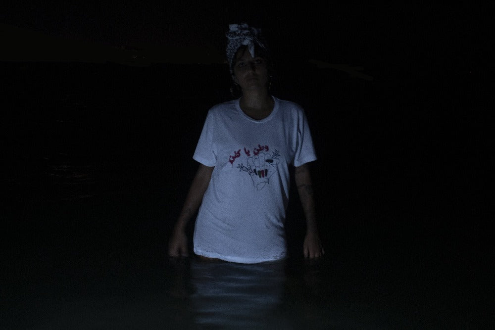 Woman wearing a white shirt standing in water. Shirt has a woman's fist, holding barb wire, bleeding, nails painted black, red and green,  a tattoo of Afghanistan on the wrist, with the words "watan ya kafan" written in Dari in blood above. 