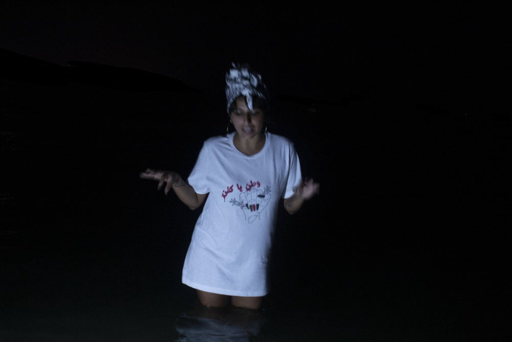Woman wearing a white shirt standing in water. Shirt has a fist drawn on it, and the words "watan ya kafan" in Dari. The woman looks away from the camera, down at the water. 