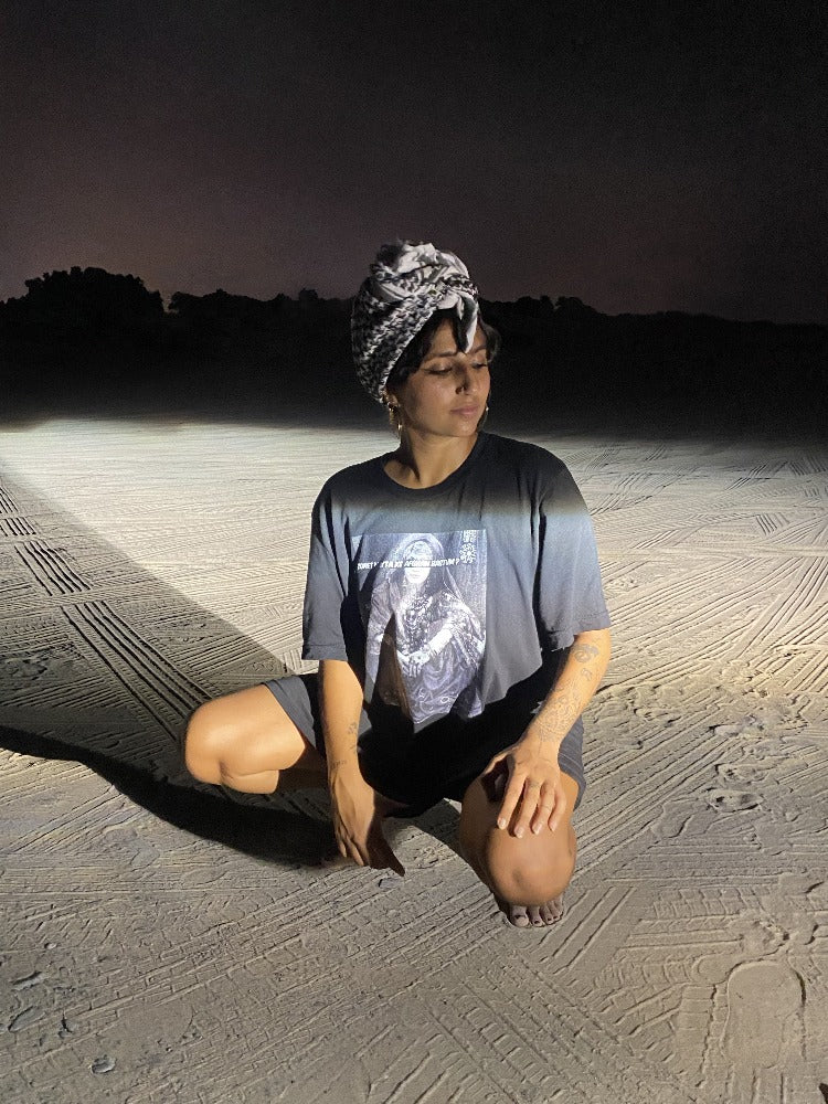 A woman kneels on the beach in a black shirt, with a Palestinian scarf on her head and her eyes closed. On her shirt is a woman in traditional Afghan clothes and jewellery sits looking at the camera, but her eyes are covered with the words "Zoret Mayta Ke Afghan Hastum" in Latinised Dari, in white font.