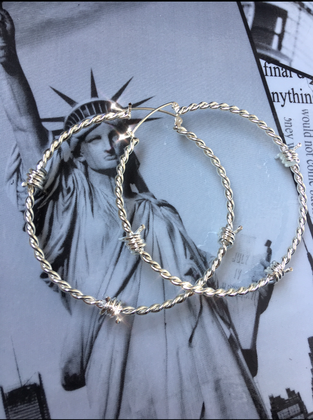 The Kabul House Essential Barb Wire Hoops - Blingistan, silver barbwire earrings on statue of liberty print.