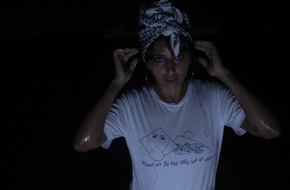 Woman in a white shirt with a kite, mountains with houses on it standing in water, with all black around and a Palestinian scarf around head, looking at the camera