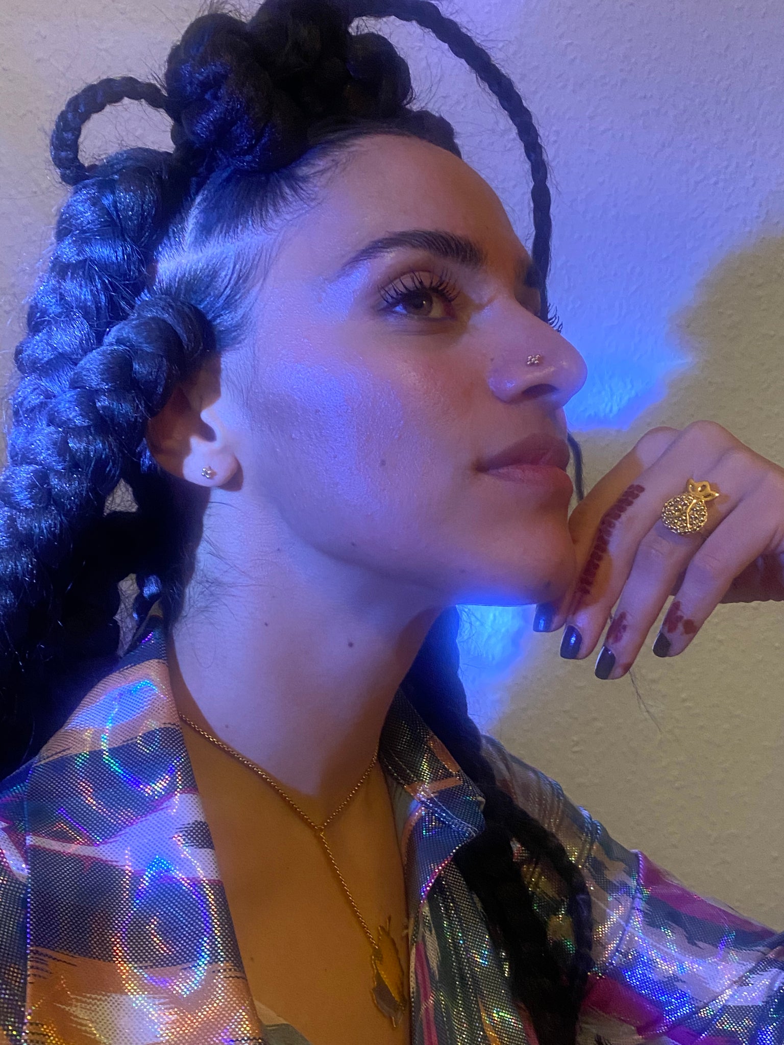 A blue ish light shines on the profile of a woman's face wearing gold studs in her nose and ear, and a pomegranate shaped gold ring.