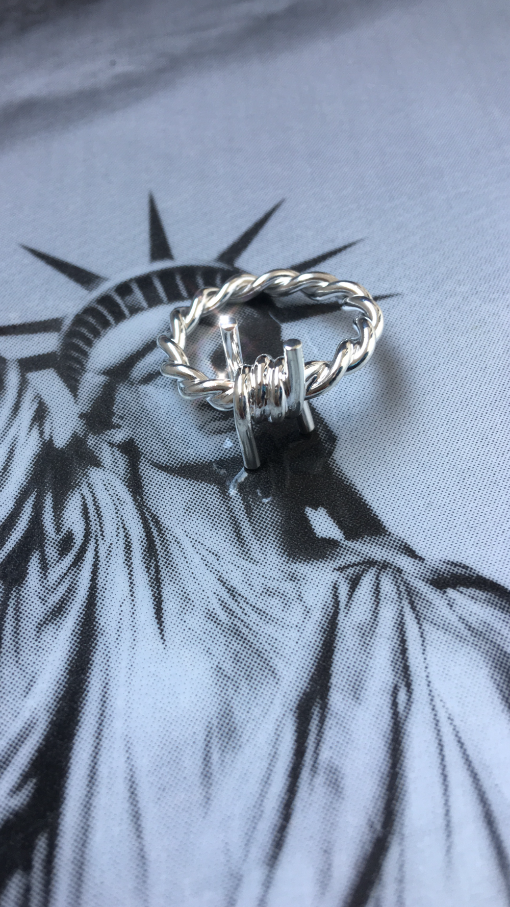 The Kabul House Essential Barb Wire Ring - Blingistan, silver barbwire ring on print of statue of liberty.