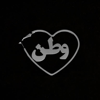 Silver hoops in the shape of a heart with the word 'watan' on it against a black back drop.