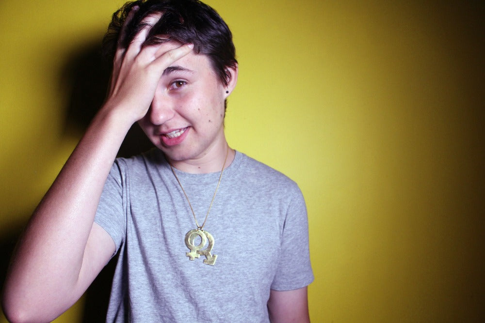 The Pride and Patriarchy Male & Female Necklace - Blingistan, person standing in front fo yellow backdrop in grey shirt wearing gold plated symbolic necklace has their hand on their head moving their hair.