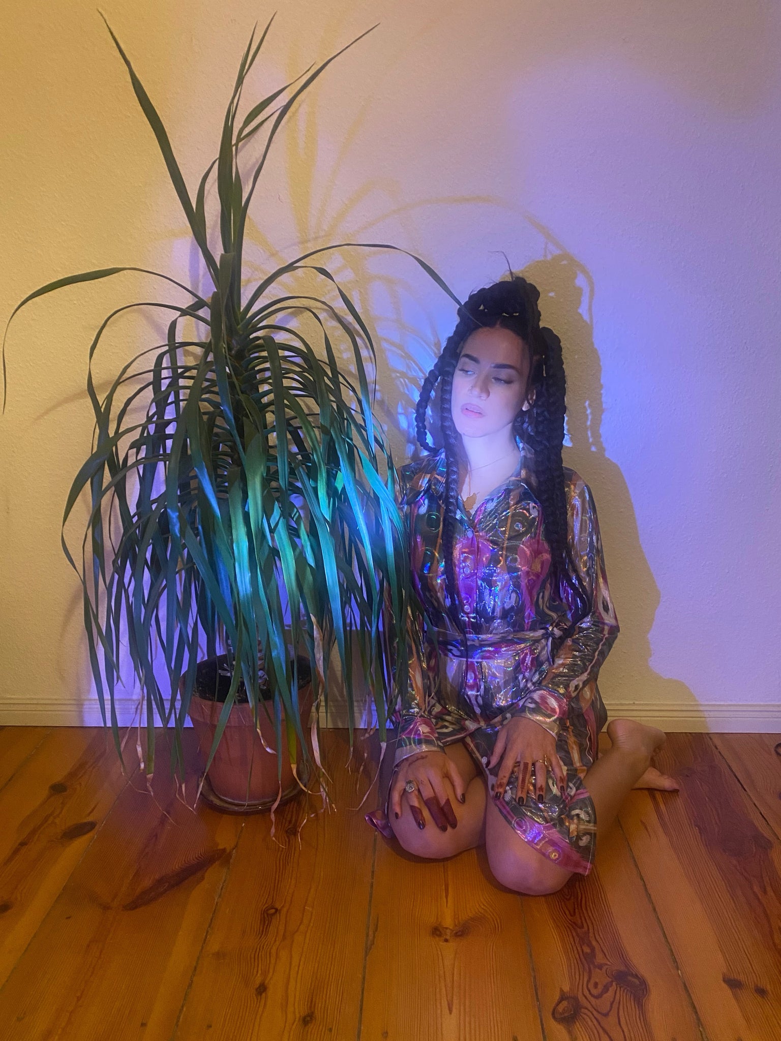 A woman sits next to a plant, wearing a holographic psychadelic ikat dress.